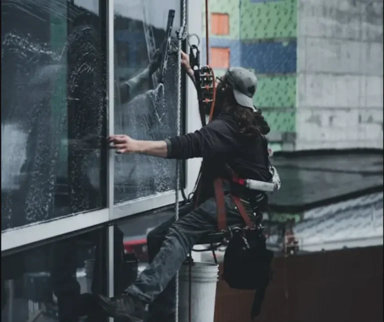 Professional commercial window cleaner suspended on ropes cleaning a high-rise building window as part of seasonal exterior maintenance.